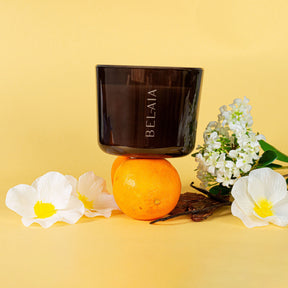 Bougie naturelle parfumée monoï imparfaite 200h sur un fond jaune et posée sur une orange entourée de fleurs de monoi 