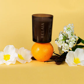 Bougie naturelle parfumée monoï imparfaite 70h en verre marron foncé avec une gravure Belaia sur un fond jaune et posée sur une orange entourée de fleurs de monoi 