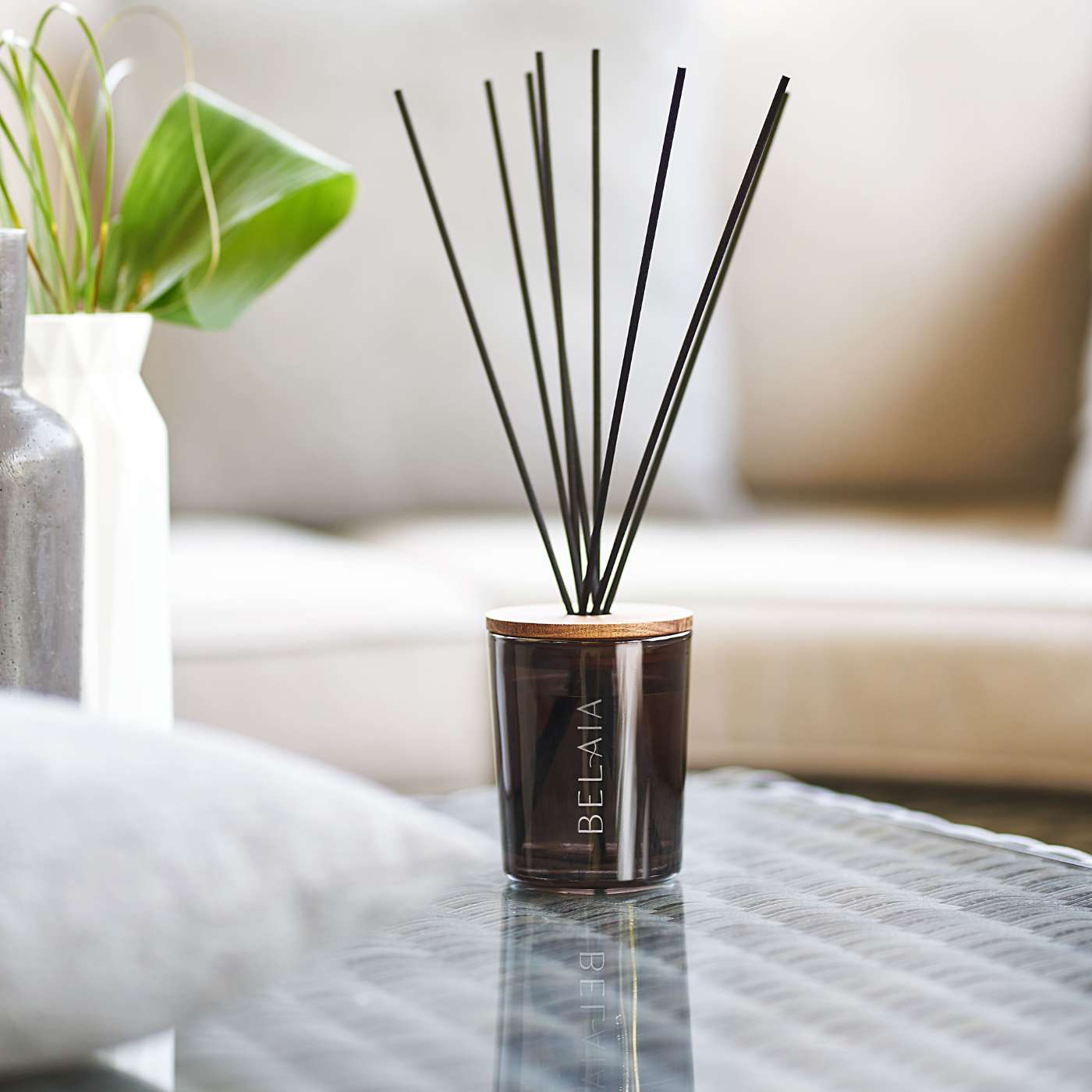 Diffuseur de parfum dans un verre marron foncé avec une gravure au nom de la marque Belaia avec des bâtonnets posé sur une table avec en fond d'image une plante à gauche et un canapé beige qui apporte une touche chaleureuse