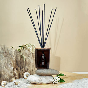 Diffuseur de parfum rechargeable en verre foncé marron avec une gravure Belaia imparfait parfumée Sable Chaud 150ml posée sur des galets entouré de coquillages 